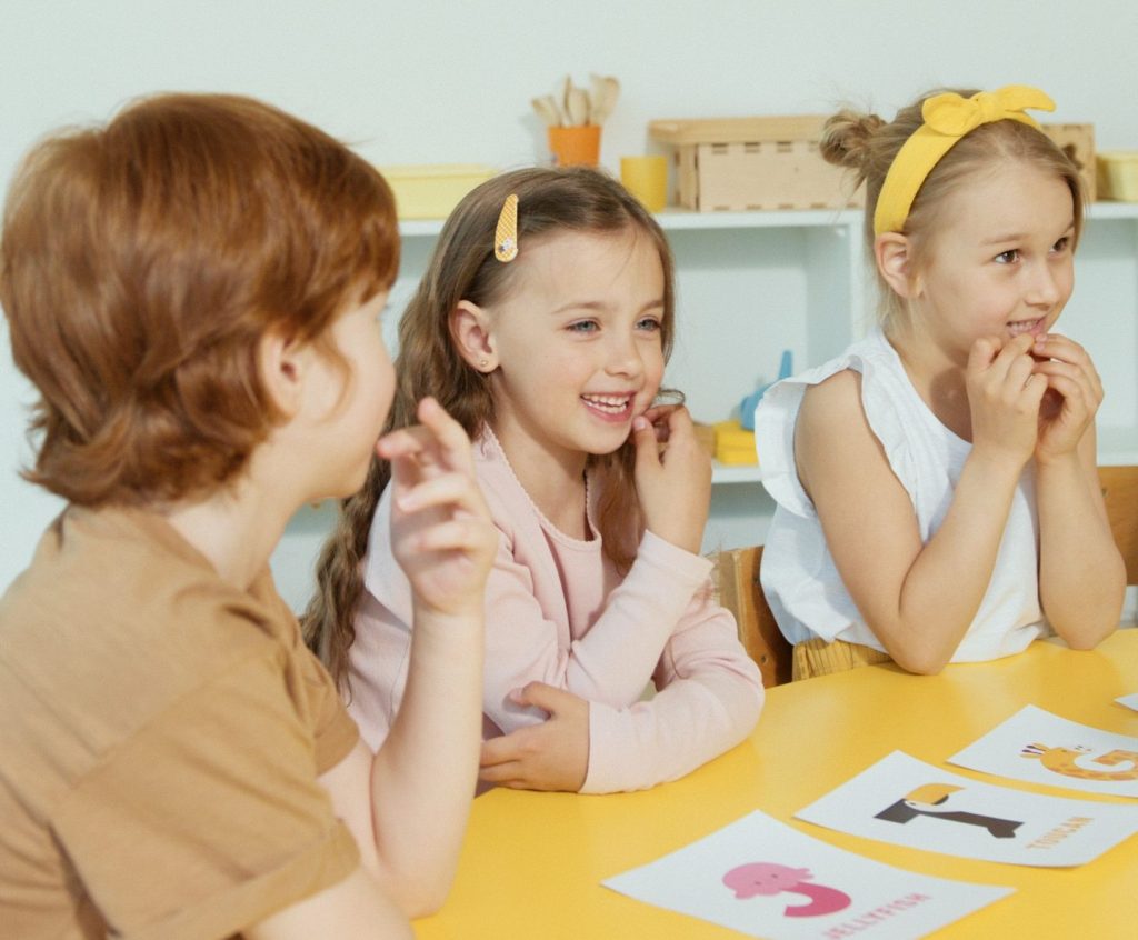 Group of children laughing and participating in a classroom setting around a yellow table.